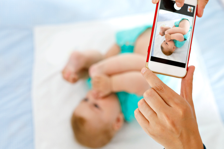 Parent taking photo of a baby with smartphone. Adorable newborn child taking foot in mouth